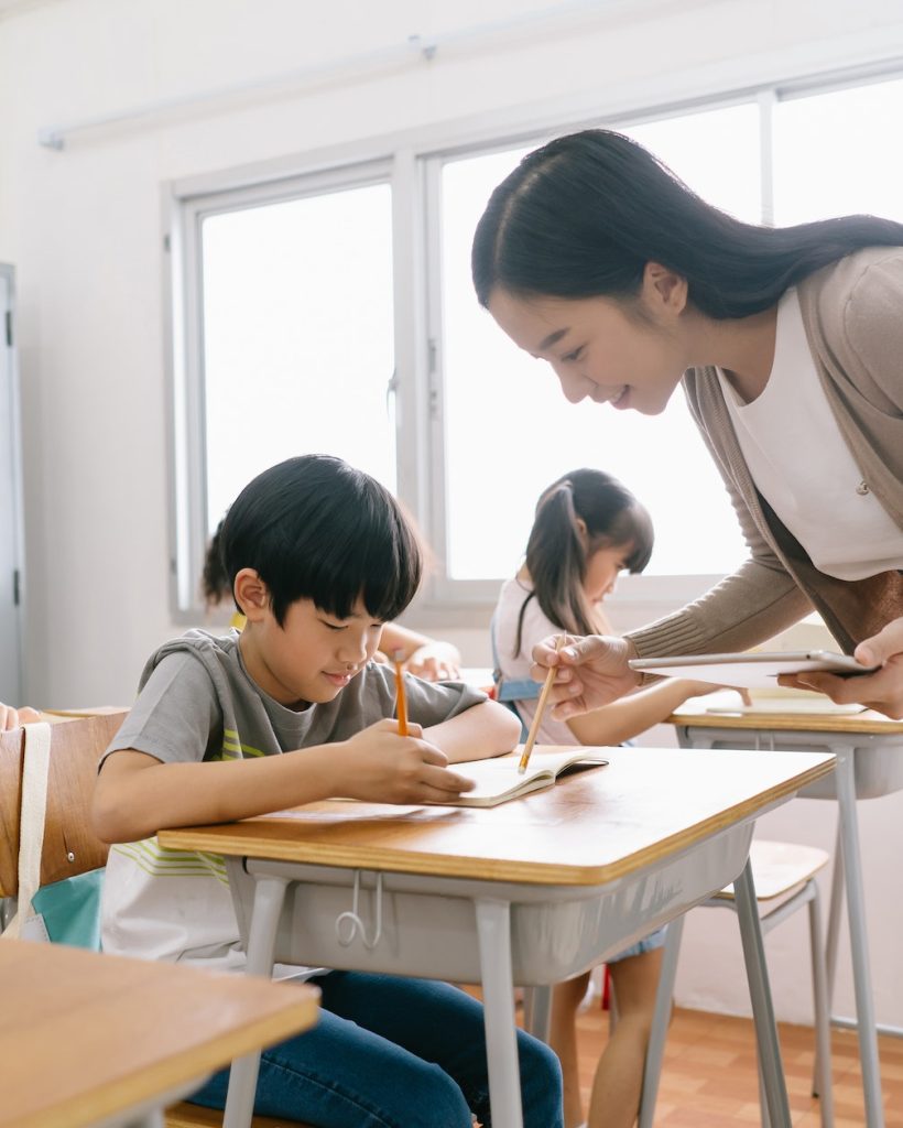 Portrait of Asian female teacher helping elementary school boy in classroom at school.